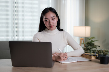 An attractive and happy young Asian woman is working on her laptop at a table in her living room. Working from home, freelance, online meeting