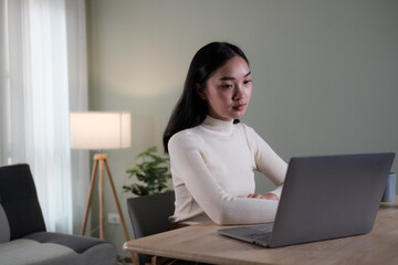 An attractive and happy young Asian woman is working on her laptop at a table in her living room. Working from home, freelance, online meeting