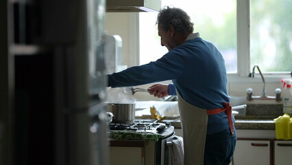 Candid senior man cooking food standing by kitchen stove stirring ingredients with utensil. Elderly...