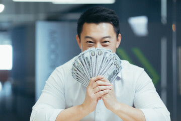 Close-up photo of a smiling young Asian man in a white shirt, sitting in the office in front of the camera and holding a fan of cash money and dollar, bills in front of his face
