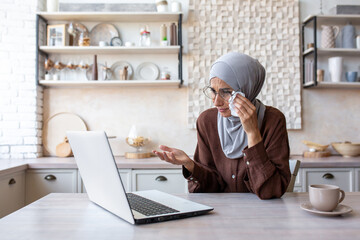 Muslim woman in hijab sitting in kitchen at home and crying in front of laptop monitor. Wipes tears with a napkin.