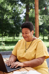 Obraz premium Vertical portrait of Black senior woman using laptop outdoors at terrace and smiling looking at computer screen