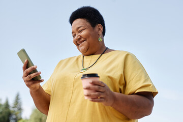 Waist up portrait of smiling Black senior woman using smartphone outdoors against blue sky, copy space