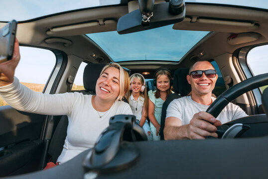 Happy young couple with two daughters inside car during auto trop. They are smiling, laughing and taking selfies using smartphone. Family values, traveling, social media and new technology concepts.