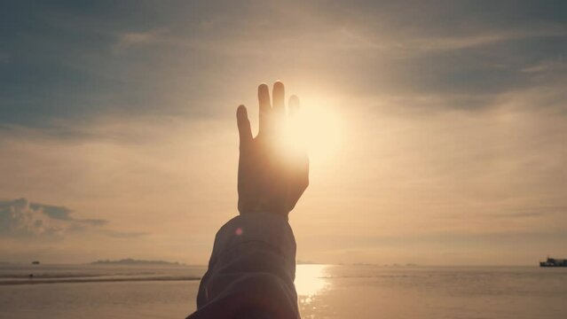 A woman's hand against the background of the summer sky. Close-up of a chewy dreamer stretching out her hand to the sun at sunset. The sun's rays shine through the hand. Summer dreaming concept.