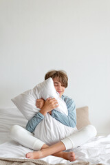 Good sleeping, time to sleep, youth daily health habit concept. Teenager boy sitting on bed, embracing pillow and smiling. Empty white wall background