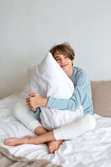 Handsome sincere teenager boy sitting on bed, embracing pillow, looking on camera. Good morning, healthy daily sleep concept, lifestyle. Minimalist neutral bedding