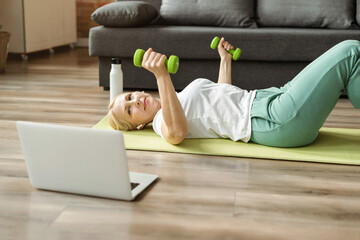 Mature woman doing fitness exercise on mat with dumbbells and watching tutorial training videos...