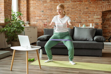 Senior woman during her fitness workout at home with rubber resistance band. She watching tutorial...