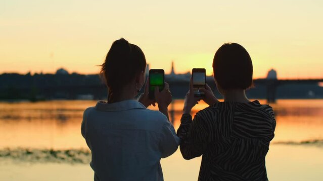 Two women take photos of river with mobile phone. Beautiful girls taking video of sunset on smartphone, female couple, friends relaxing on the beach