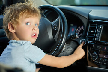 Blue-eyed boy is sitting in the driver's seat inside the car, pressed the ignition button with his...