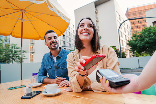 Happy couple paying the bill of the coffee shop using a cellphone contactless payment application. Young adult customer woman doing a purchase on a restaurant with a contactless smartphone banking app
