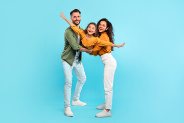 Excited parents playing with daughter holding her in arms posing on blue background, girl spreading...