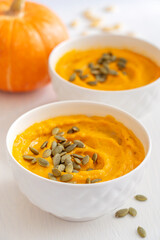 Closeup view of homemade vegetarian savory pumpkin soup made of pureed vegetables is common Thanksgiving dish decorated with seeds served in bowls on white wooden table with ingredients for dinner
