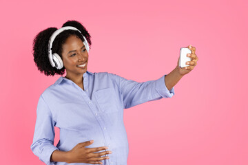 Glad millennial black lady with belly in wireless headphones listens to music, enjoys free time, makes selfie on phone