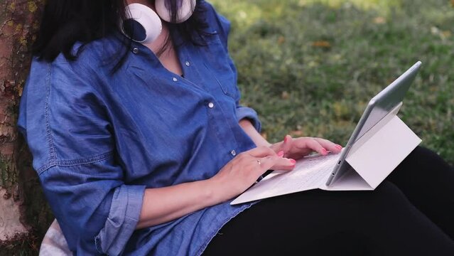 Brunette lady in casual clothes typing text on tablet sitting near tree in city garden woman with headphones working on internet via device on lawn grass