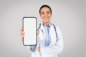 Smiling brazilian female doctor showing smartphone with blank screen at camera, standing on grey...