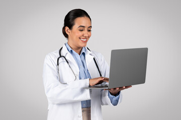 Smiling confident latin female doctor in white coat with stethoscope typing on laptop isolated on gray background
