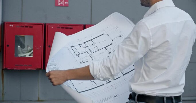 A man in a white shirt unfolds a large paper drawing in front of him, the man stands against the background of red fire shields