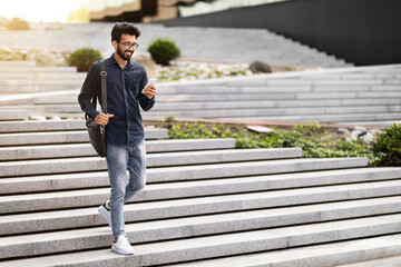 Cheerful indian guy going down stairs outdoors, using cell phone