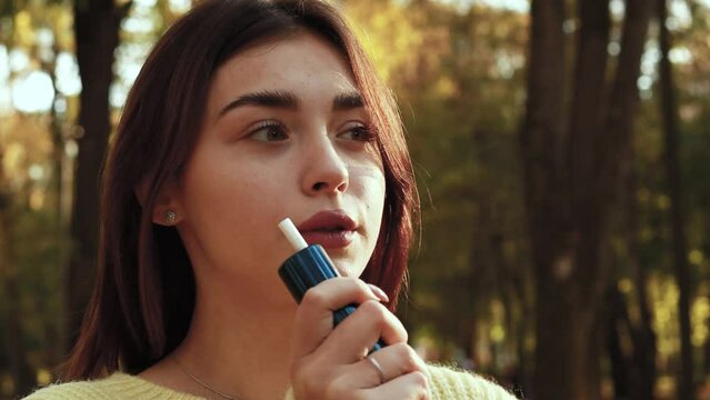 Slow motion footage. Young woman in warm sunny autumn park season smoking tobacco device electronic cigarette heater vape. Smoke and steam system, fall, yellow orange red leaves
