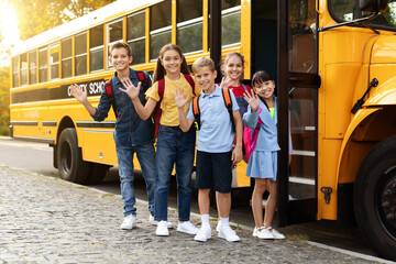 Group Of Happy Children Standing Near School Bus And Waving At Camera