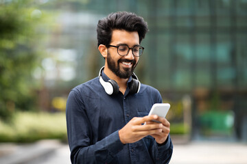 Handsome young indian guy walking by street with phone, headphones