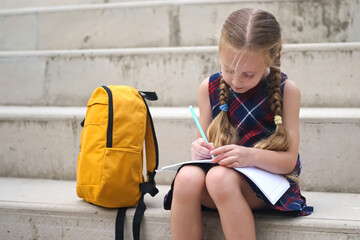 Cheerful pupil girl with a nearby yellow backpack, engrossed in outdoor homework tasks after...