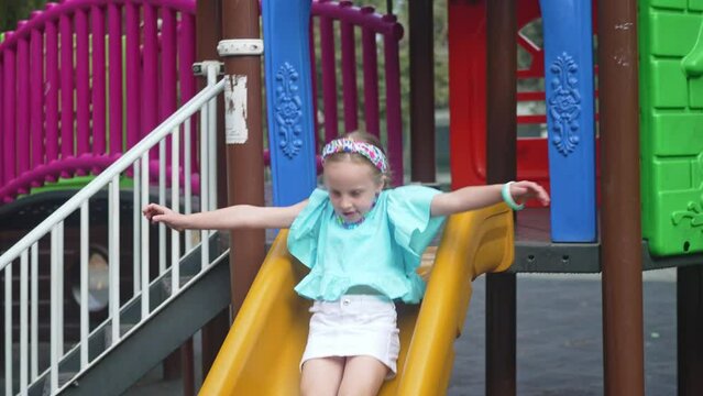 Playful little girl in casual blue clothes rides slide in maze on children playground smiling schoolgirl plays attraction in public park child enjoys weekend