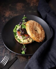 Vegetable bagel with green leaves, onions, tomatoes and hummus in a black dish with a black napkin and black background.