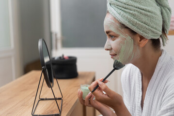 Smiling woman applying clay mask on face with brush looking in table mirror lady with towel on head...