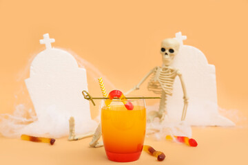 Glass of delicious cocktail for Halloween with candies and skeleton on beige background