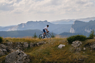 Professional male cyclist is riding a gravel bike on a gravel road with an amazing view of the mountains. Cyclist is practicing on gravel road. Man is riding on top of a hill.Bucegi Mountains, Romania - obrazy, fototapety, plakaty
