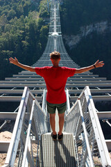 A male tourist is enjoying a trip to the mountains, standing on a suspension bridge with his arms...