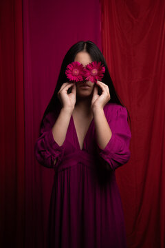 art portrait of woman in pink dress holding red germini flowers in front of her eyes,
