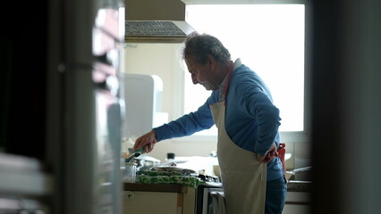 Naklejka na ściany i meble Retired Chef in Apron Focused on Home Cooking, candid senior man preparing food by kitchen stove