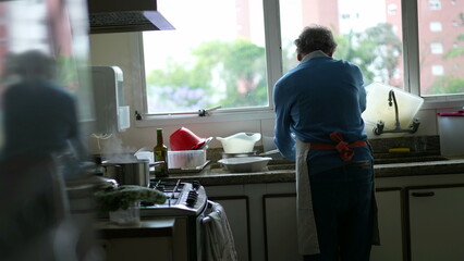 Senior by kitchen sink washing dishes and preparing food at stove. Elderly person cooking food wearing apron, candid genuine retired lifestyle - obrazy, fototapety, plakaty