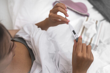 Young woman looks at electronic thermometer with high temperature closeup female with flu rests sitting in bed in light bedroom upper view symptom of disease