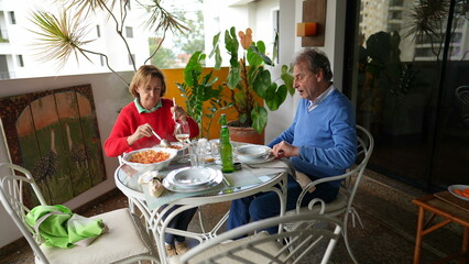 Senior couple sitting down at apartment terrace for lunch. Married elderly husband and wife in balcony serving food, domestic lifestyle scene in old age retirement years