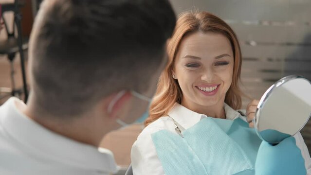 Smiling and happy woman in dental clinic satisfied with the result of dentist's work. The dentist shows the patient the condition of her oral cavity with the help mirror. Looking at herself in mirror