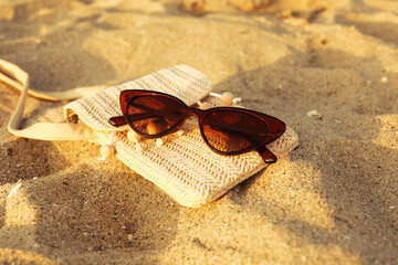 Stylish brown sunglasses and wicker bag on sand