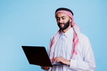 Muslim man wearing traditional thobe and ghutra headscarf holding laptop and broswing social media. Arab person dressed in islamic clothes using portable computer and checking internet page