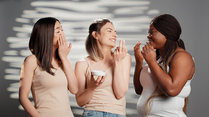 Group of beautiful girls applying moisturizing cream on camera, promoting natural skincare products in studio. Diverse women using moisturizer and serum for new beauty campaign.