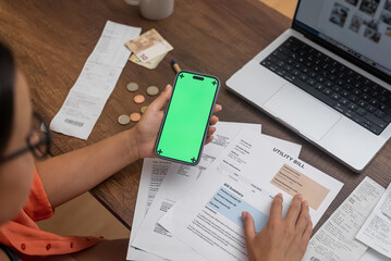 Fototapeta na wymiar Lady holds smartphone with green chromakey screen for posting image sitting at table with documents of utility bills woman counts money to repay loan via device