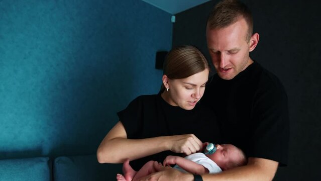 Caucasian parents wearing black t-shirts stand in the room. Happy couple holding their newborn baby.