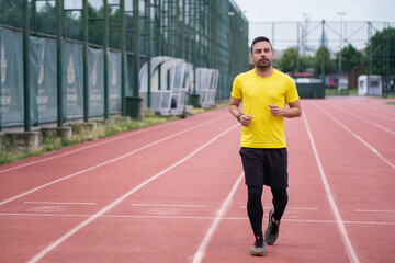 Young man in bright t-shirt running along empty track with red rubber surface and white markings at...