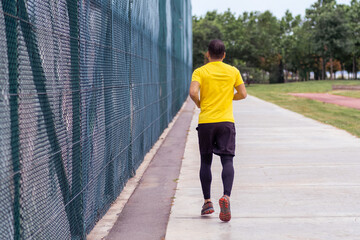 Sportsman in tracksuit running along empty track by high grid barrier on urban sports ground in green park activities of healthy lifestyle backside view