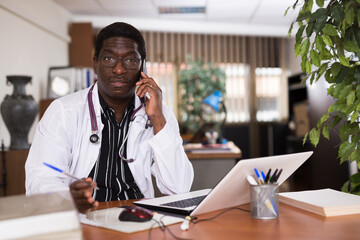 Highly qualified african american doctor conducts a patient consultation on a mobile phone, sitting at a workdesk in ..the resident's office