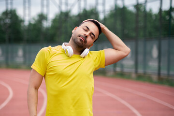 Man in yellow t-shirt doing neck side bend preparing muscles for athletic training on running track at city sports ground basic physical exercise for warming up