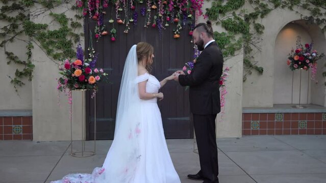 Tall dark-haired groom putting ring on hand of bride standing outdoors near the wedding venue, decorated with flowers, bouquet, ivy on walls. Profile shot. High quality 4k footage